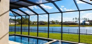 Screened pool enclosure overlooking a peaceful lake, green lawn, and waterfront homes under a bright blue sky